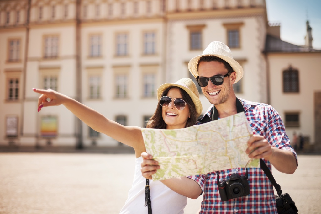 Day, summer. Young, smiling people. He is wearing a shirt with a card, she is wearing a white T-shirt. They are both wearing small hats and sunglasses. The man is holding a map in his hand and a camera around his neck. The woman is pointing at something with her finger. In the background, a historic building.